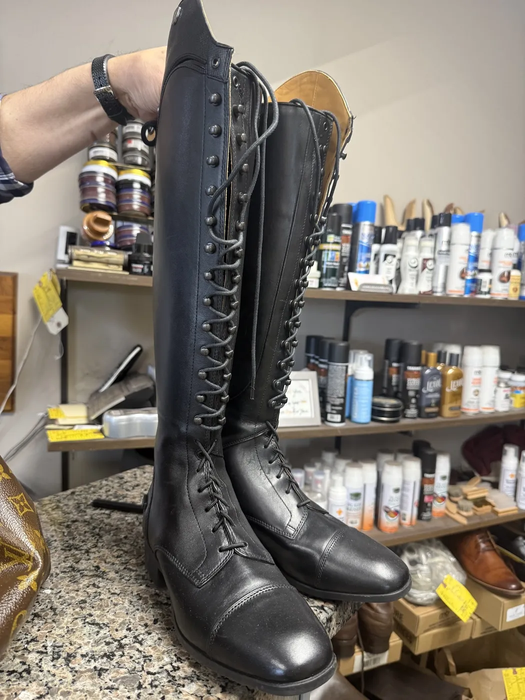 Tall black lace-up riding boots on the counter in front of shelves of shoe care products